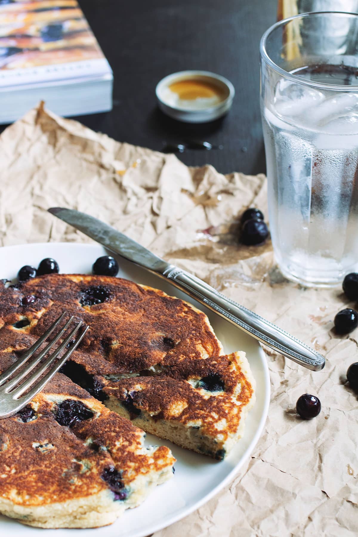 A thick blueberry pancake on a white plate with a slice taken out of it.