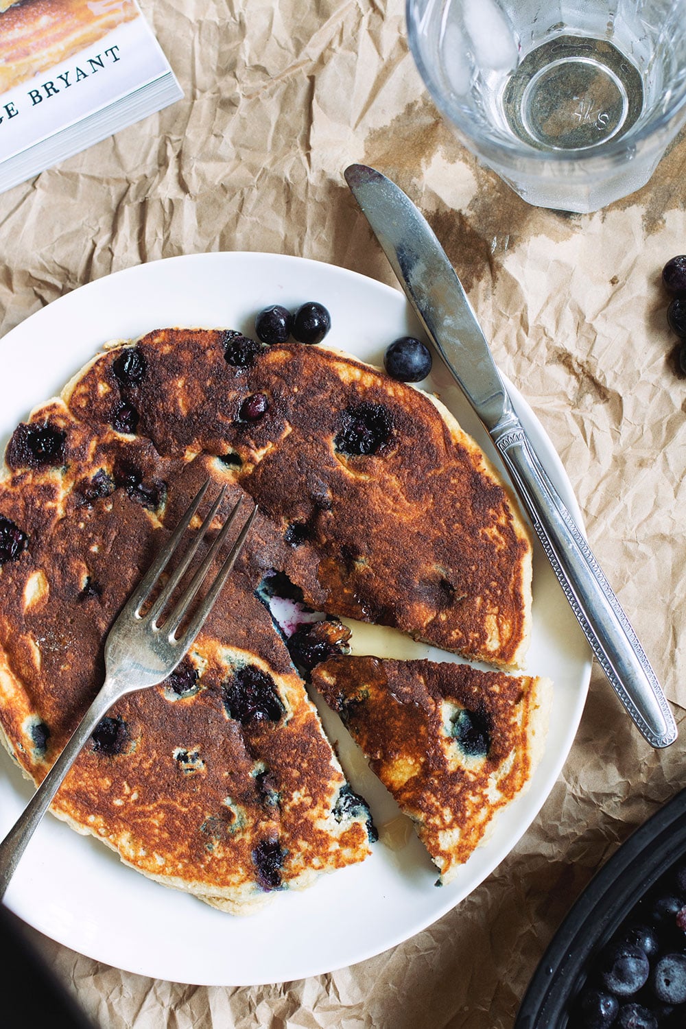 A thick blueberry pancake on a white plate with a slice taken out of it.