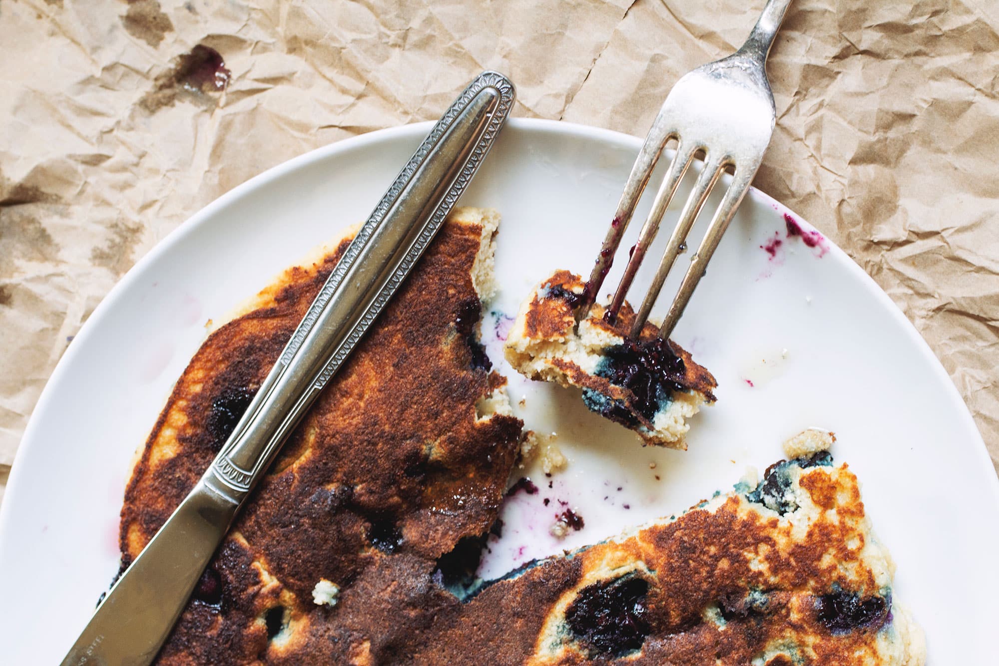 A thick blueberry pancake on a white plate with a fork taking a bite out.