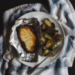 Miso-glazed cod in foil on a white plate with a pile of brussels sprouts and a fork.