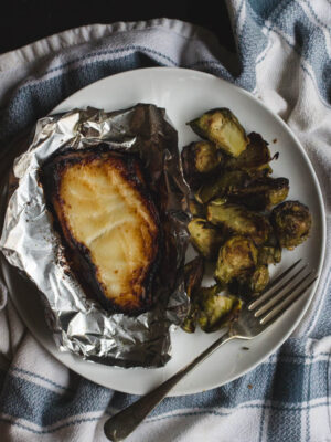 Miso-glazed cod in foil on a white plate with a pile of brussels sprouts and a fork.