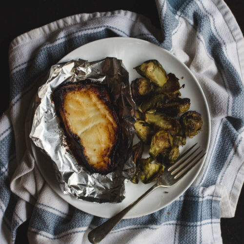Miso-glazed cod in foil on a white plate with a pile of brussels sprouts and a fork.