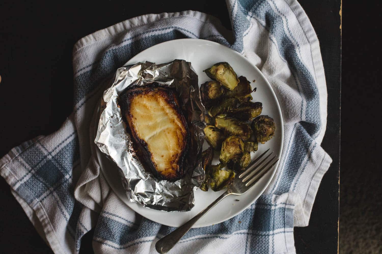 Miso-glazed cod in foil on a white plate with a pile of brussels sprouts and a fork.