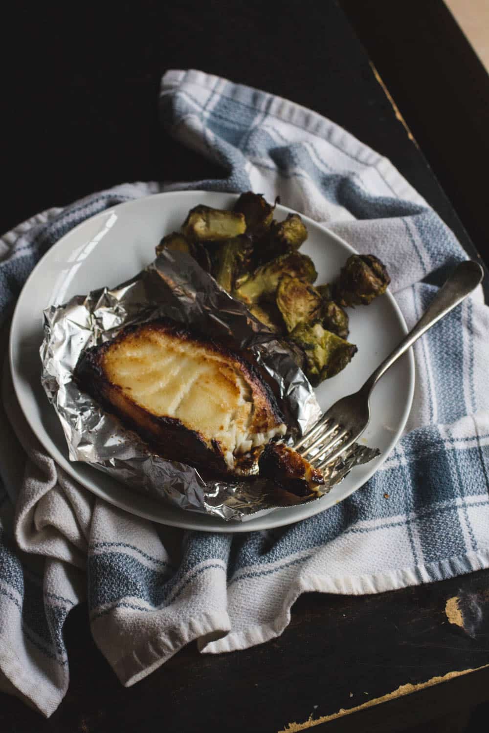 Miso-glazed cod in foil on a white plate with a pile of brussels sprouts with a fork taking a bite of fish.