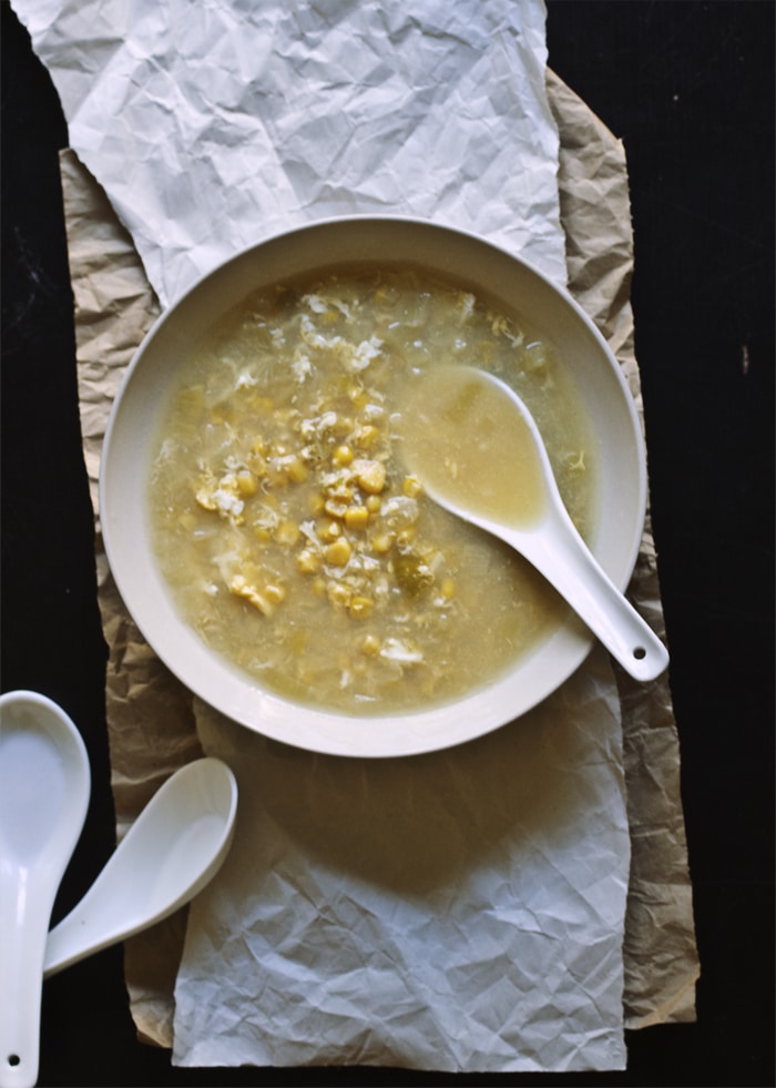 A taupe bowl full of egg drop corn soup with a soup spoon in it on top of a brown sheet of paper.