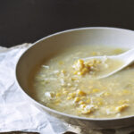 A taupe bowl full of egg drop corn soup with a soup spoon on top of a white and brown sheet of paper.