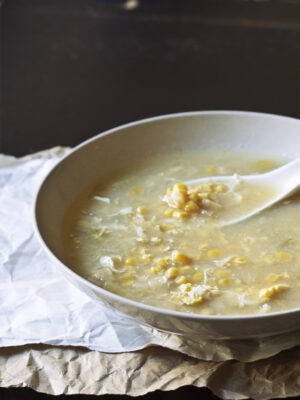 A taupe bowl full of egg drop corn soup with a soup spoon on top of a white and brown sheet of paper.
