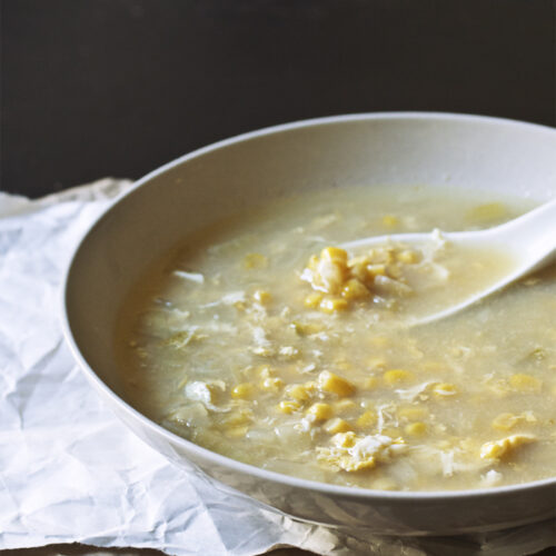 A taupe bowl full of egg drop corn soup with a soup spoon on top of a white and brown sheet of paper.