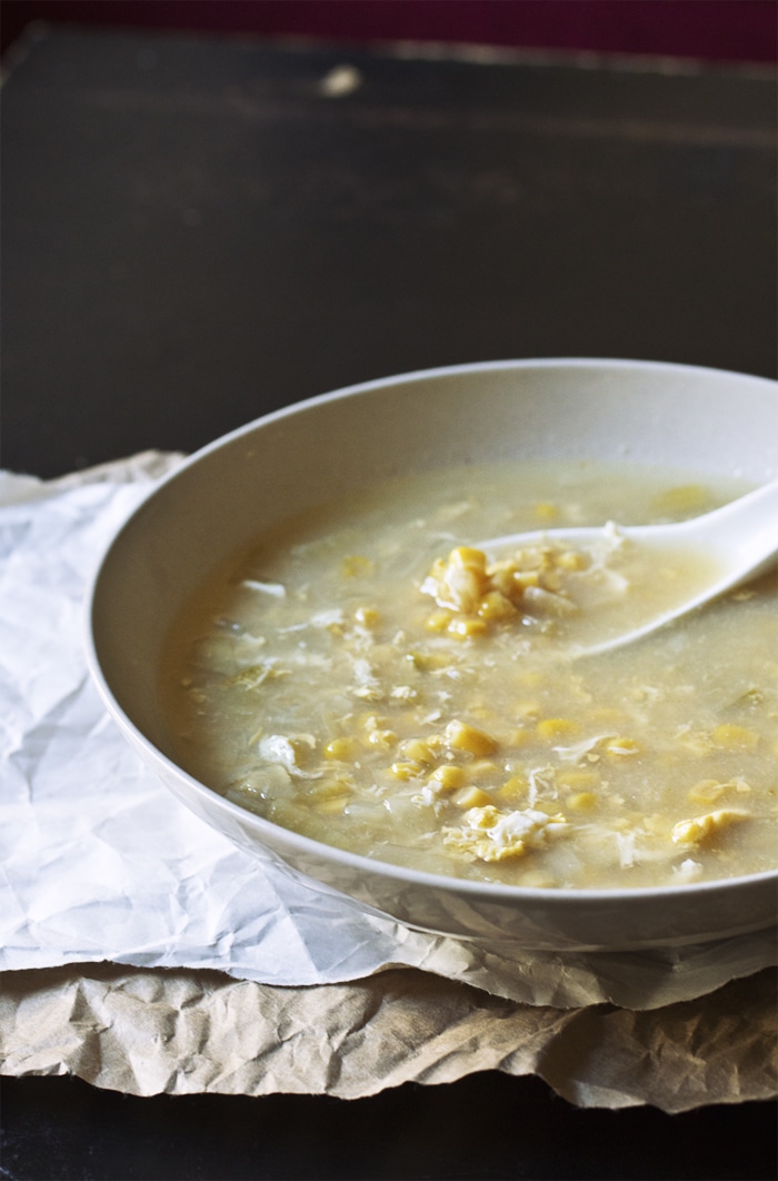 A taupe bowl full of egg drop corn soup with a soup spoon on top of a white and brown sheet of paper.