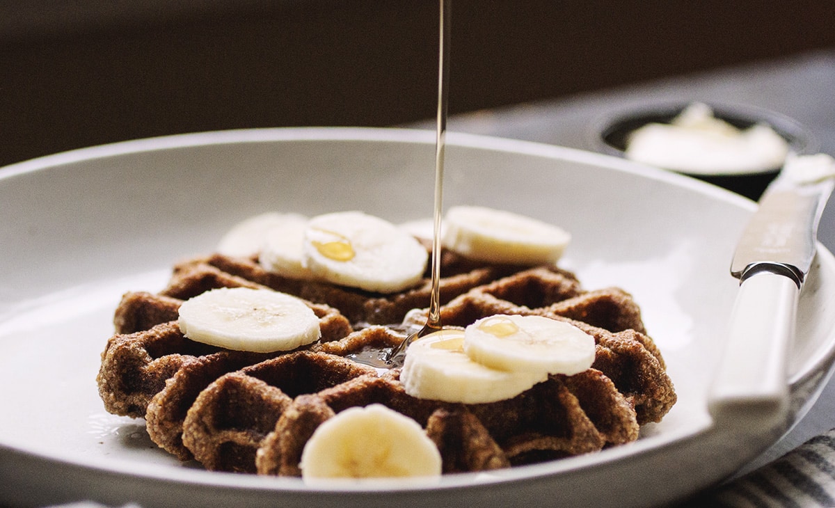 Close up on syrup being poured on a waffle topped with banana slices on a white plate.