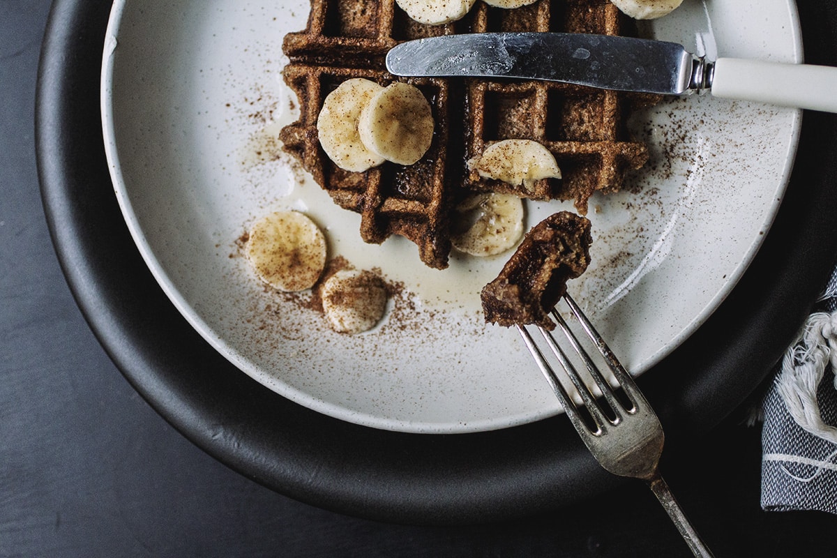 A fork taking a bite of a waffle topped with banana slices on a white plate.