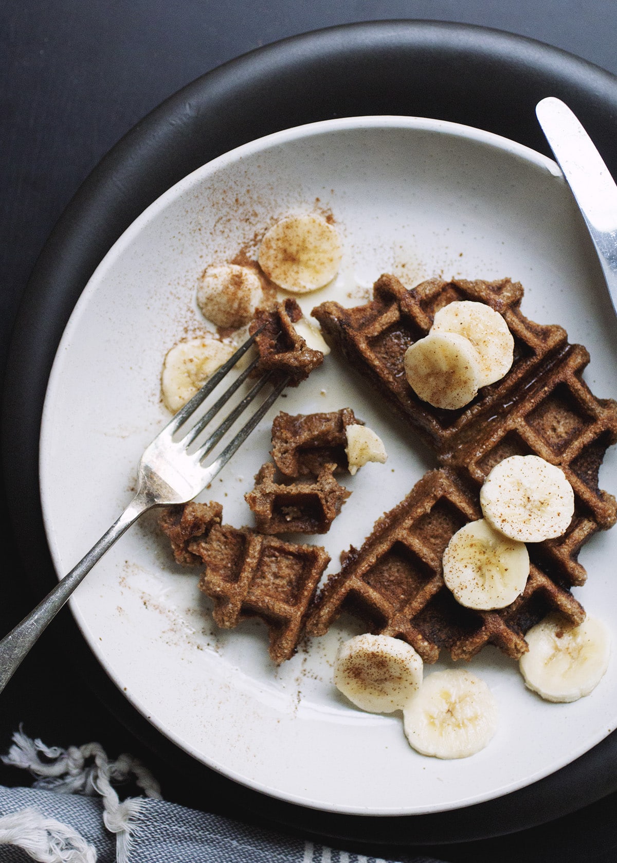 A buckwheat waffle with banana slices and a fork on a white plate.
