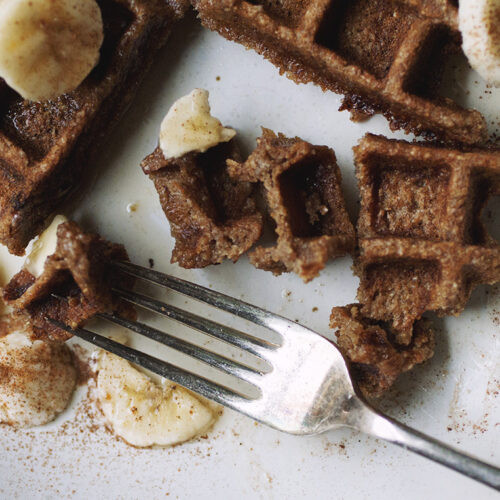 Pieces of a waffle with banana slices and a fork on a white plate.