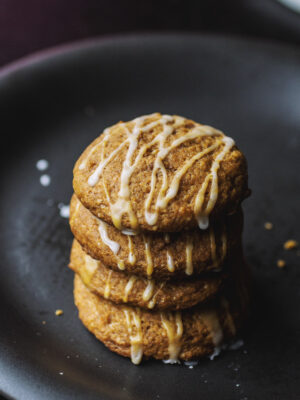 A stack of four pumpkin cookies drizzled with a white glaze on a black plate.