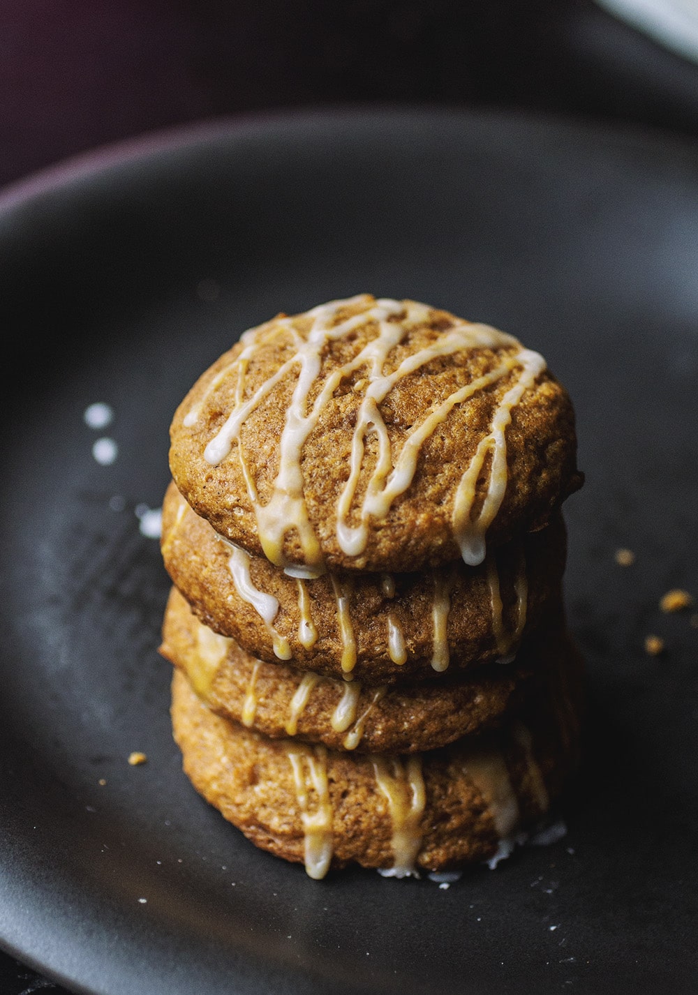 A stack of four pumpkin cookies drizzled with a white glaze on a black plate.