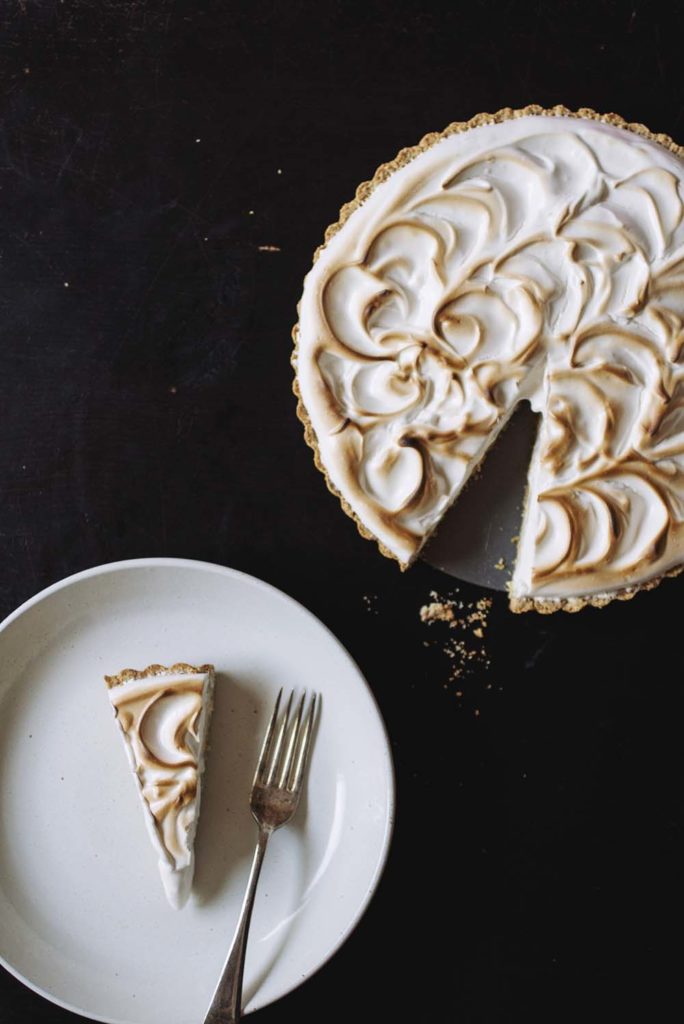 A slice of lemon meringue pie on a white plate next to the whole tart missing one slice.