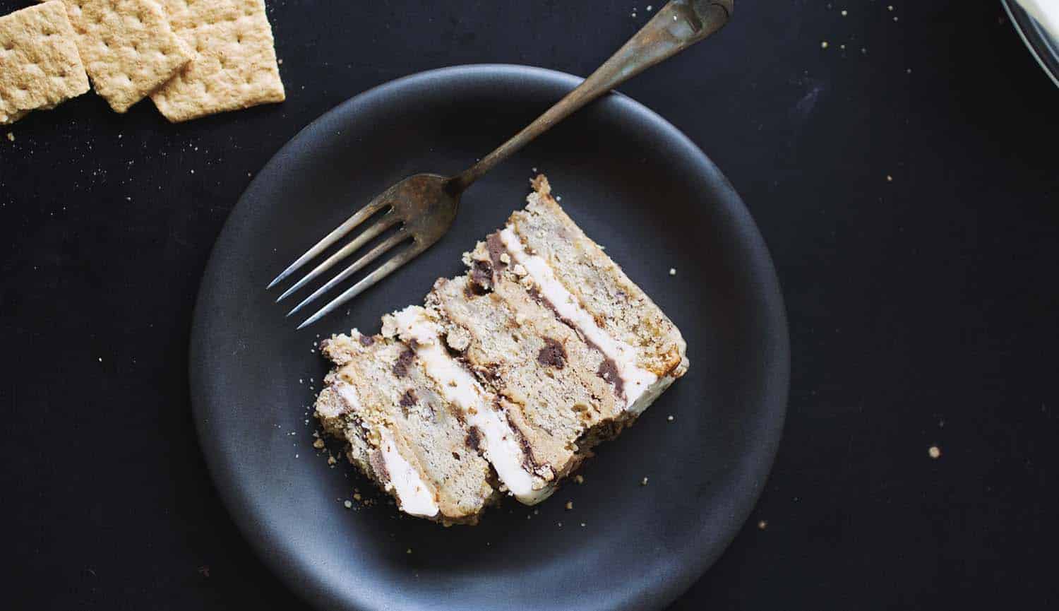 A slice of s\'mores cake layered with marshmallow fluff, graham frosting and chocolate ganache on a black plate with a fork.