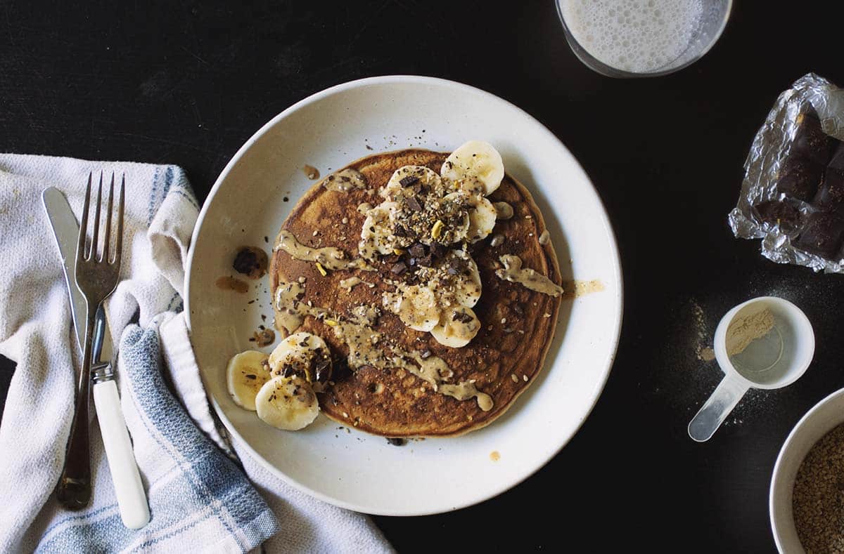 A golden brown pancake topped with sliced banana and peanut butter on a white plate next to a fork laid on top of a white towel.