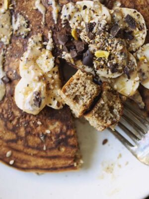 A golden brown pancake topped with sliced banana and peanut butter with a fork taking a bite.