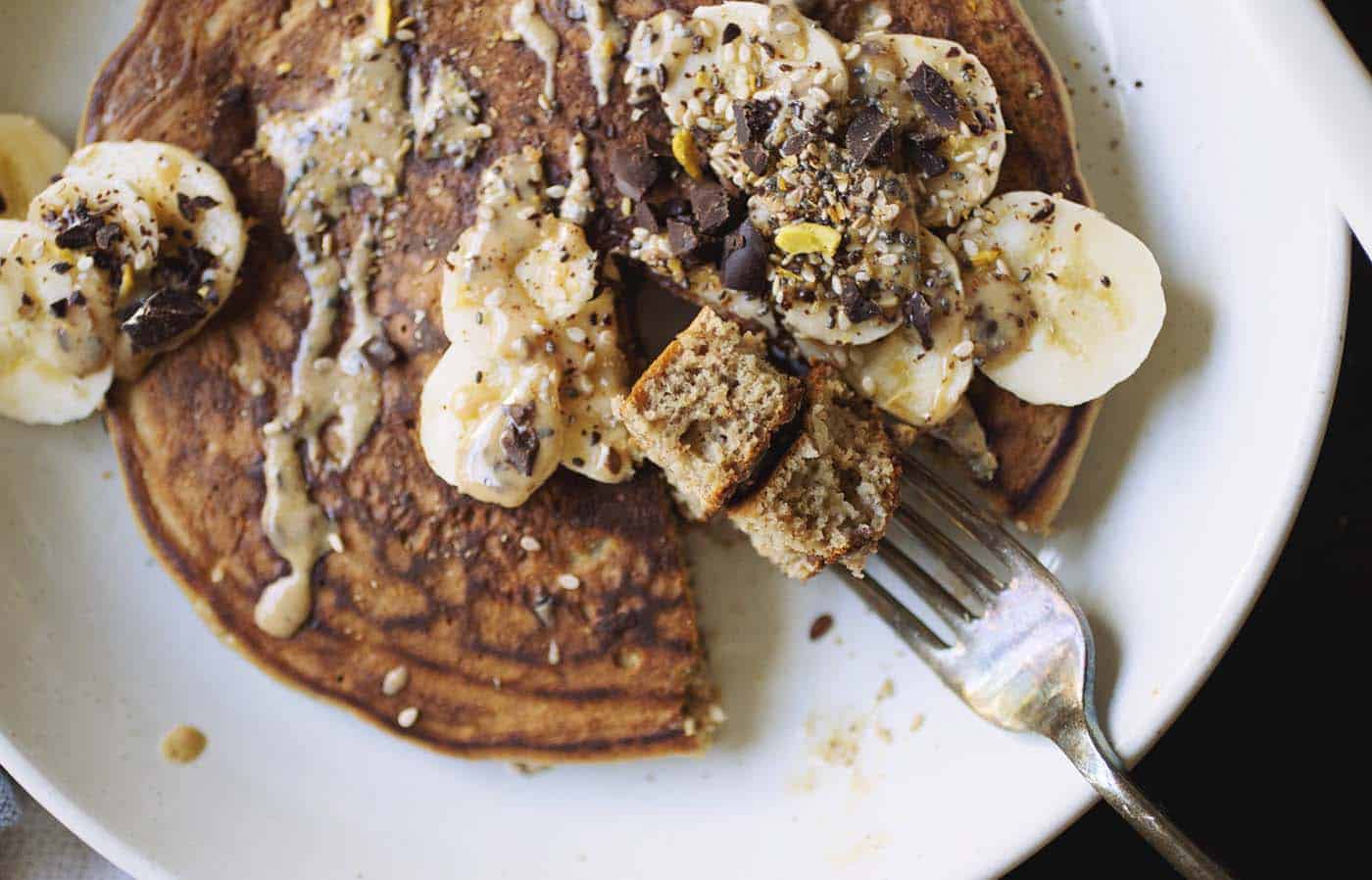 A golden brown pancake topped with sliced banana and peanut butter with a fork taking a bite.