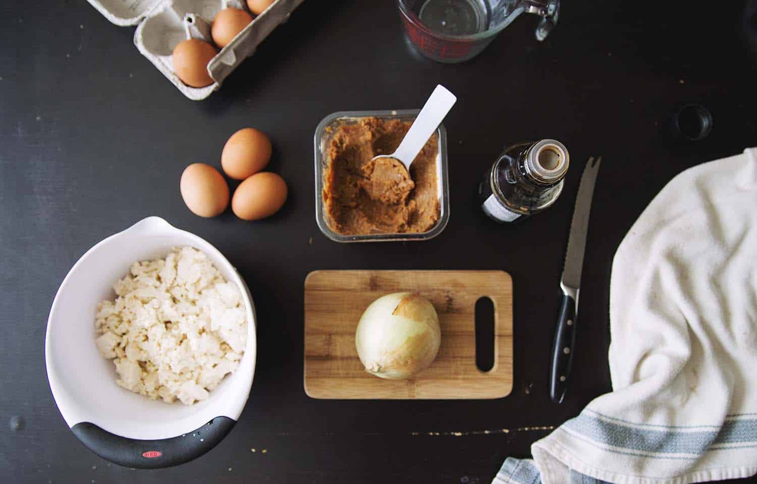A black counter with a container of miso, a bowl of crumbled tofu, a few whole eggs and an onion on a cutting board.