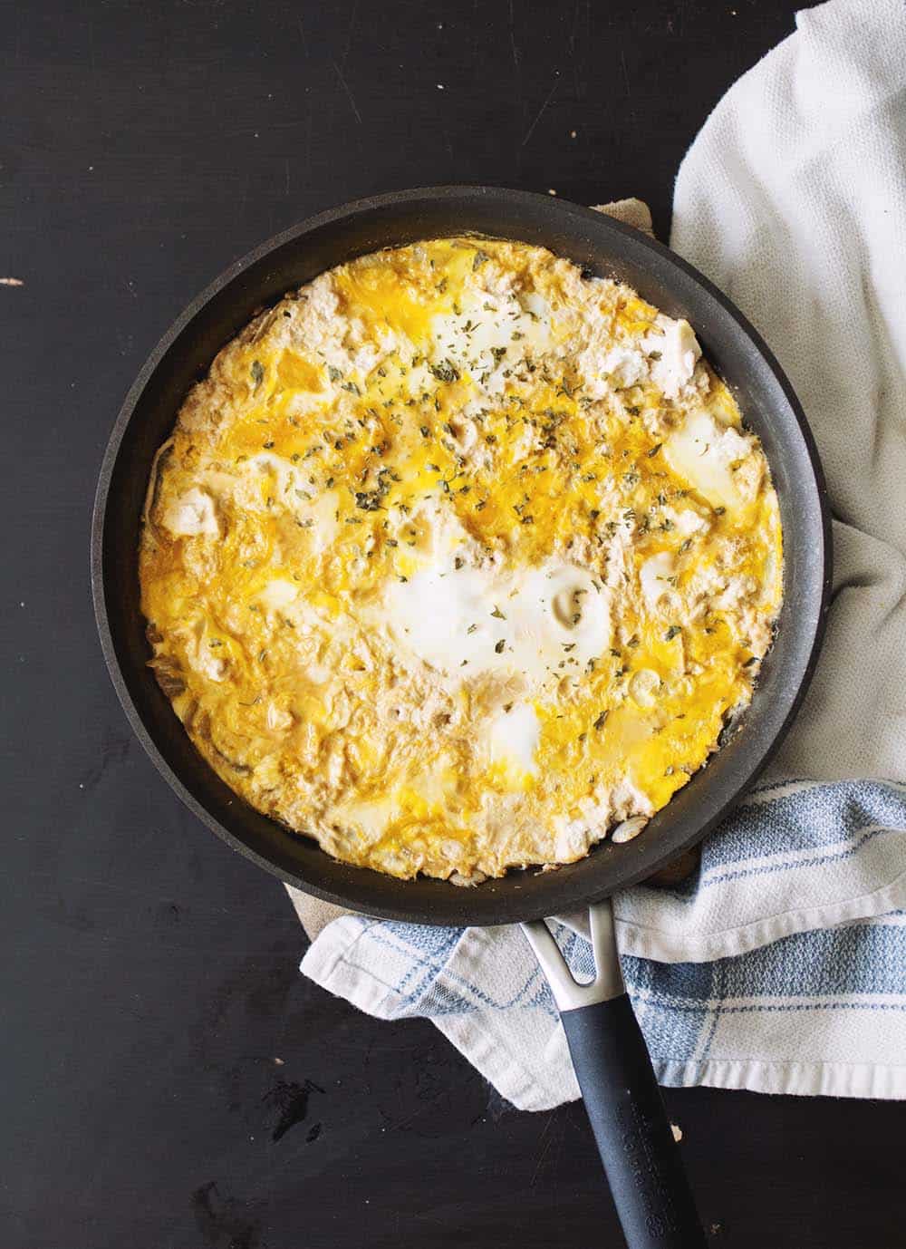 A black pan filled with a mixture of eggs, tofu and onions on a black table with a blue and white towel.