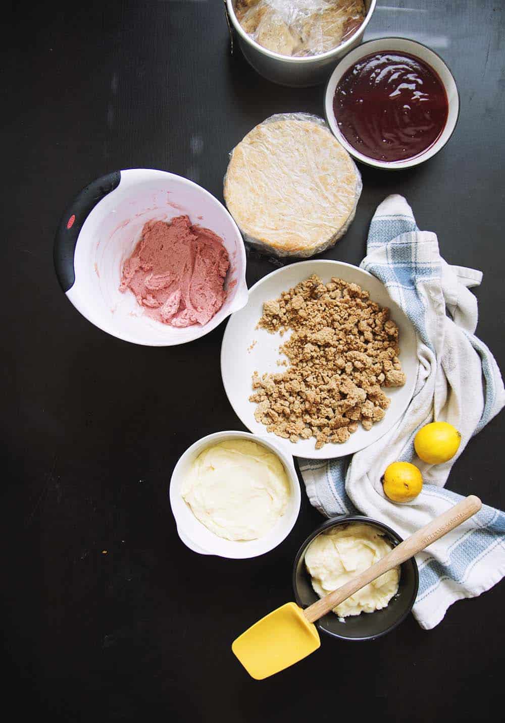A wrapped layer of cake next to a bowl of strawberry frosting, a bowl of jam, a plate full of milk crumbles, and bowls of lemon curd.