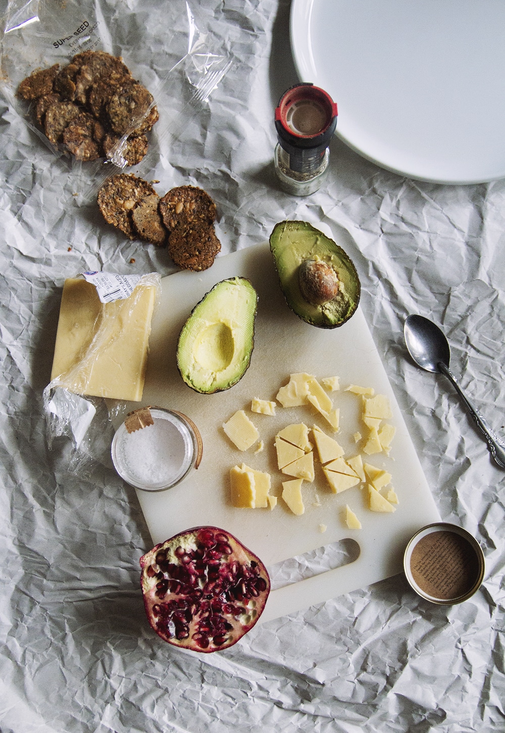 A cutting board with a block of cheese, a halved avocado, crackers and half a pomegranate.