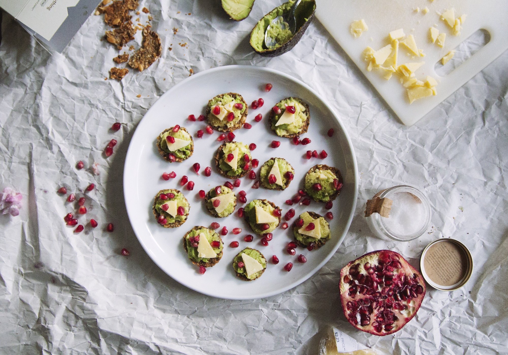 Crackers topped with avocado, cheese and pomegranate seeds on a white plate.