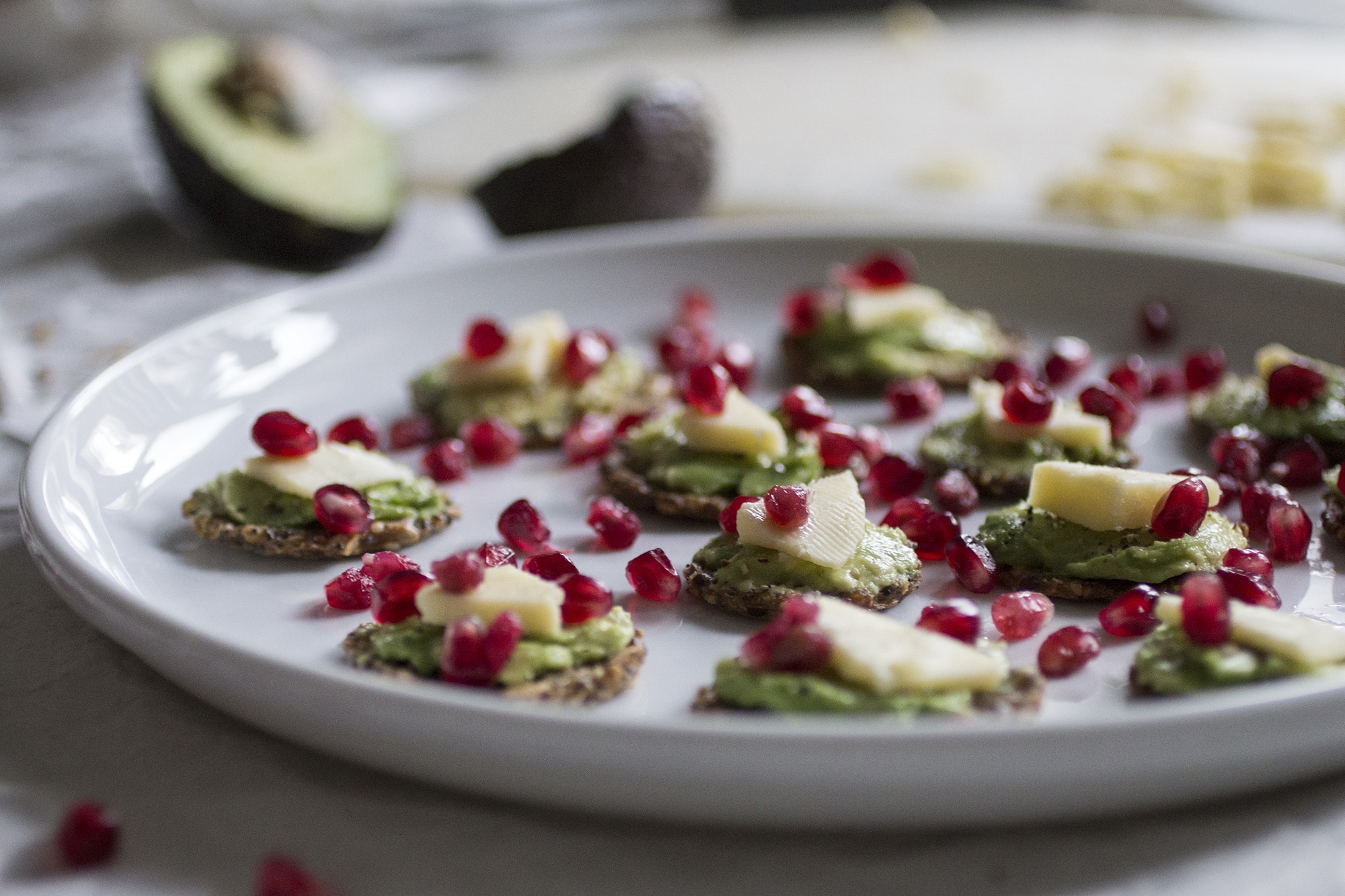 Crackers topped with avocado, cheese and pomegranate seeds on a white plate.