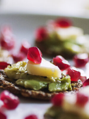A close up on a grainy cracker topped with avocado, cheese and a pomegranate seed.