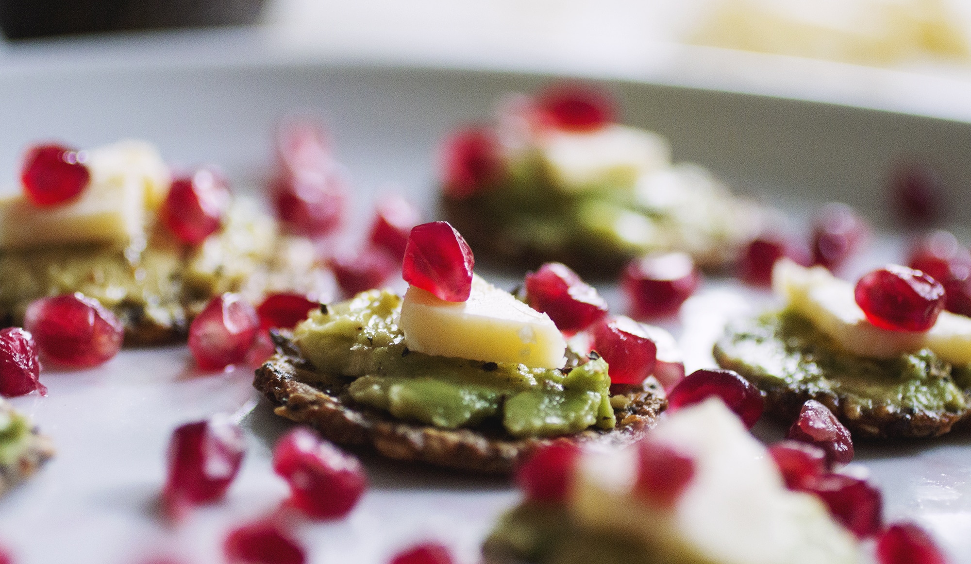 A close up on a grainy cracker topped with avocado, cheese and a pomegranate seed.