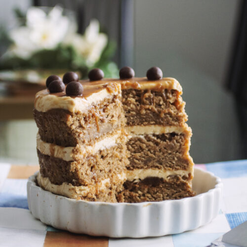 3/4 of a caramel apple cake topped with chocolate balls on a white plate with white flowers in the background.