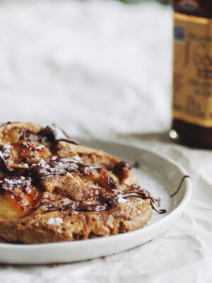 A white plate of banana buckwheat pancakes drizzled with chocolate hazelnut spread.