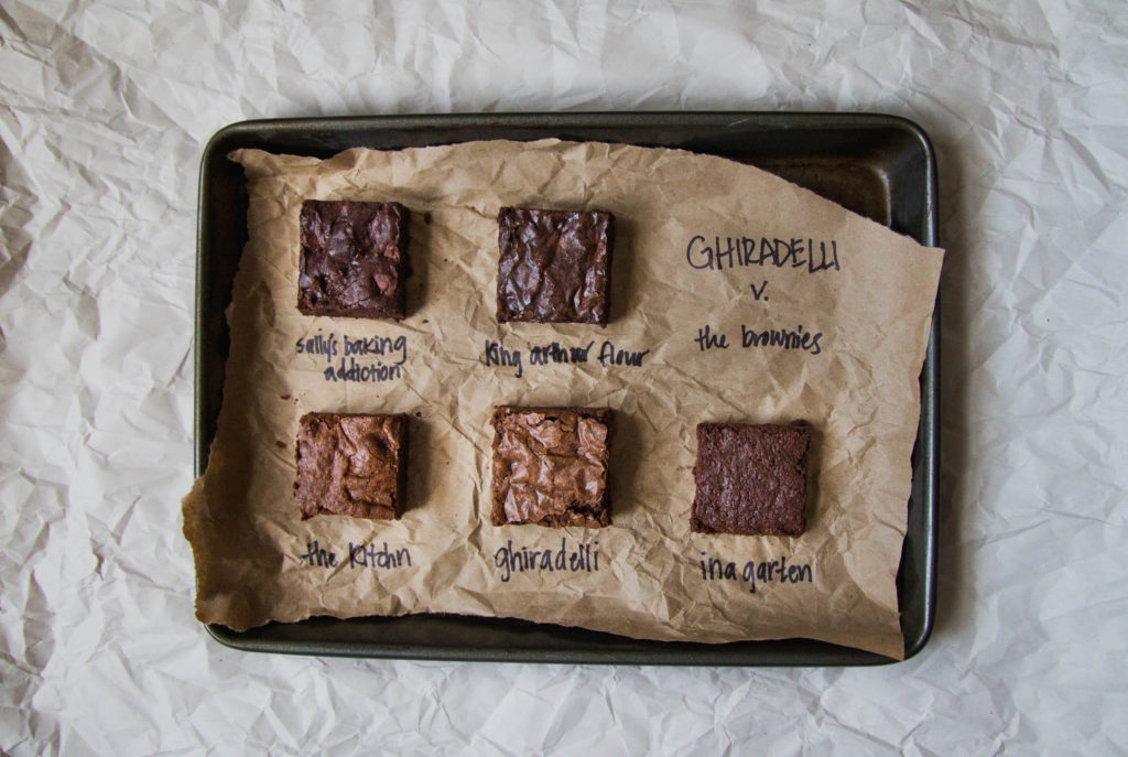 Squares of 5 different brownies on a parchment-lined baking sheet.