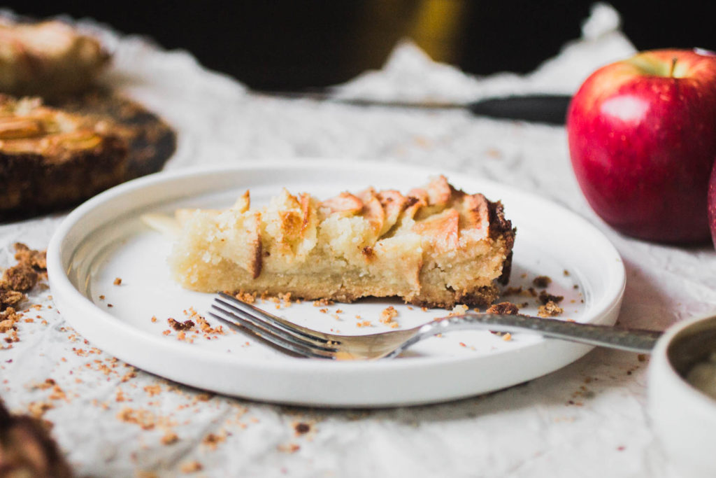 A sideways view of an apple tart with a frangipane filling on a white plate with a fork.