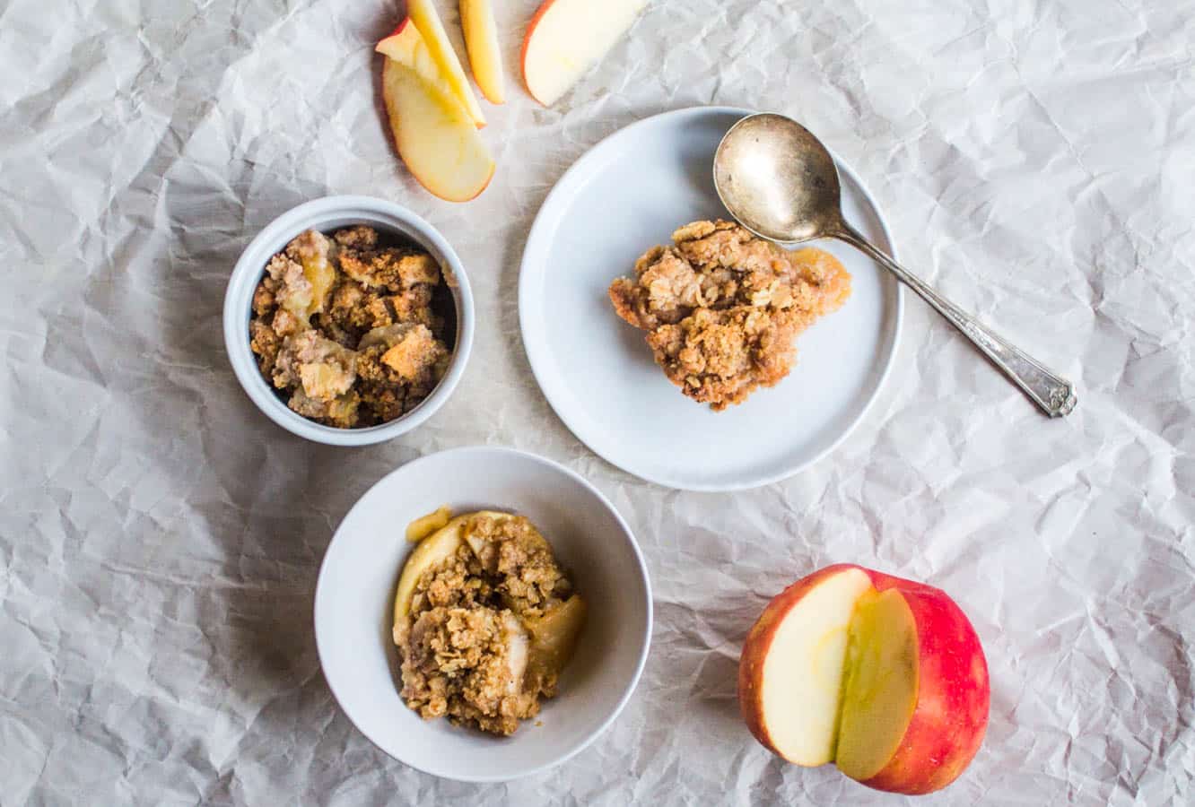 3 white bowls filled with apple crisp next to a sliced apple on a white background.