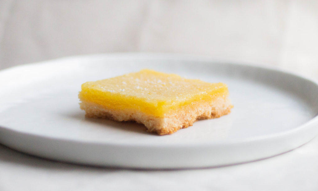 single slice of mel\'s kitchen cafe lemon bars on white plate featuring equal ratio of crust and lemon curd.