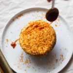 A spoon drizzling caramel on top of a coconut-topped cupcake.