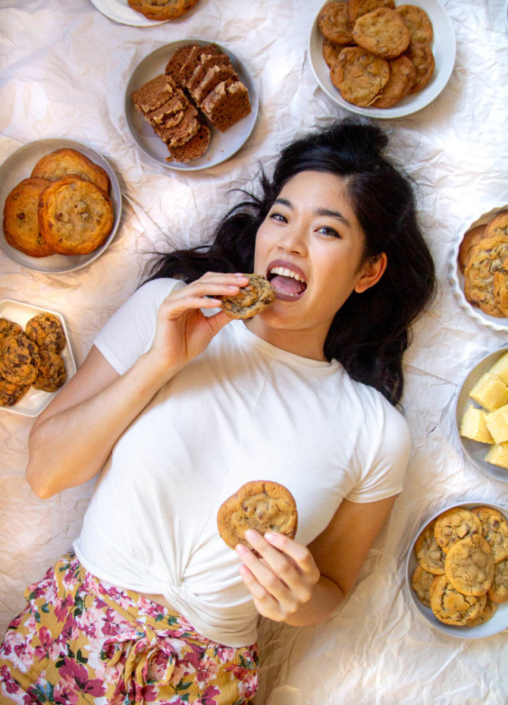 A girl smiling and holding cookies in both hands surrounded by plates of baked goods.