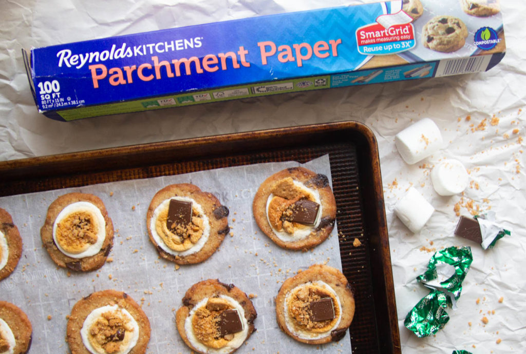 Finished s\'mores cookies on a parchment-lined baking sheet next to a roll of Reynolds Kitchens parchment paper.