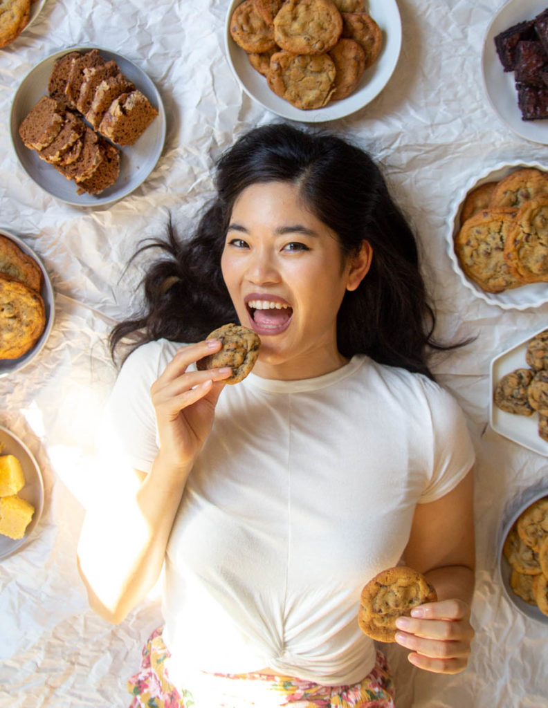A photo of The Pancake Princess smiling and holding up a cookie on a white background.