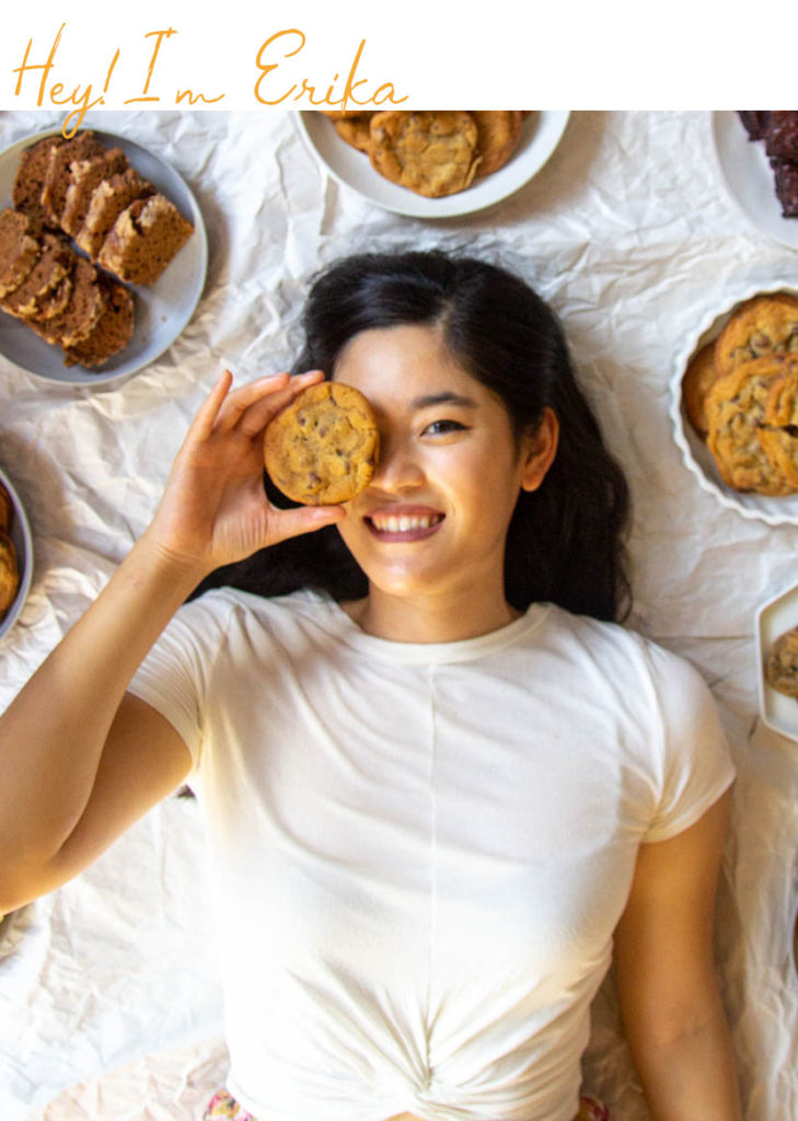 A girl surrounded by plates of baked goods holding a cookie up to her eye.