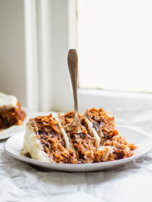 a slice of carrot cake with a fork on a white plate next to a window.