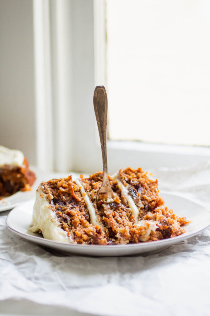 a slice of carrot cake with a fork on a white plate next to a window.