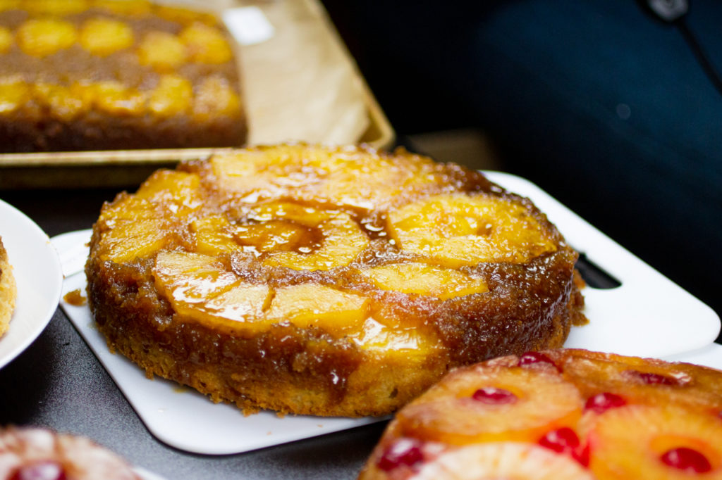 a whole pineapple upside down cake on a white cutting board near other cakes.