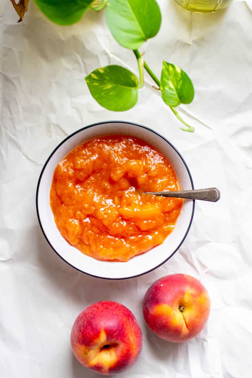 A bowl of peach compote on a white background with a couple peaches and green leaves.