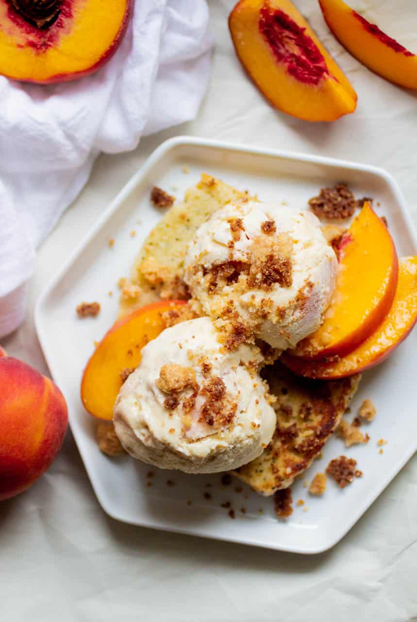 A slice of cake topped with sliced peaches, ice cream and crumbles on a white plate.