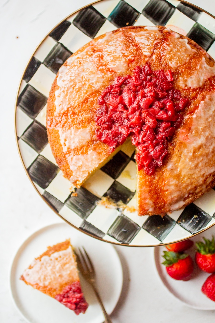 An olive oil cake with a strawberry compote with a slice missing on a black and white checkered cake plate.