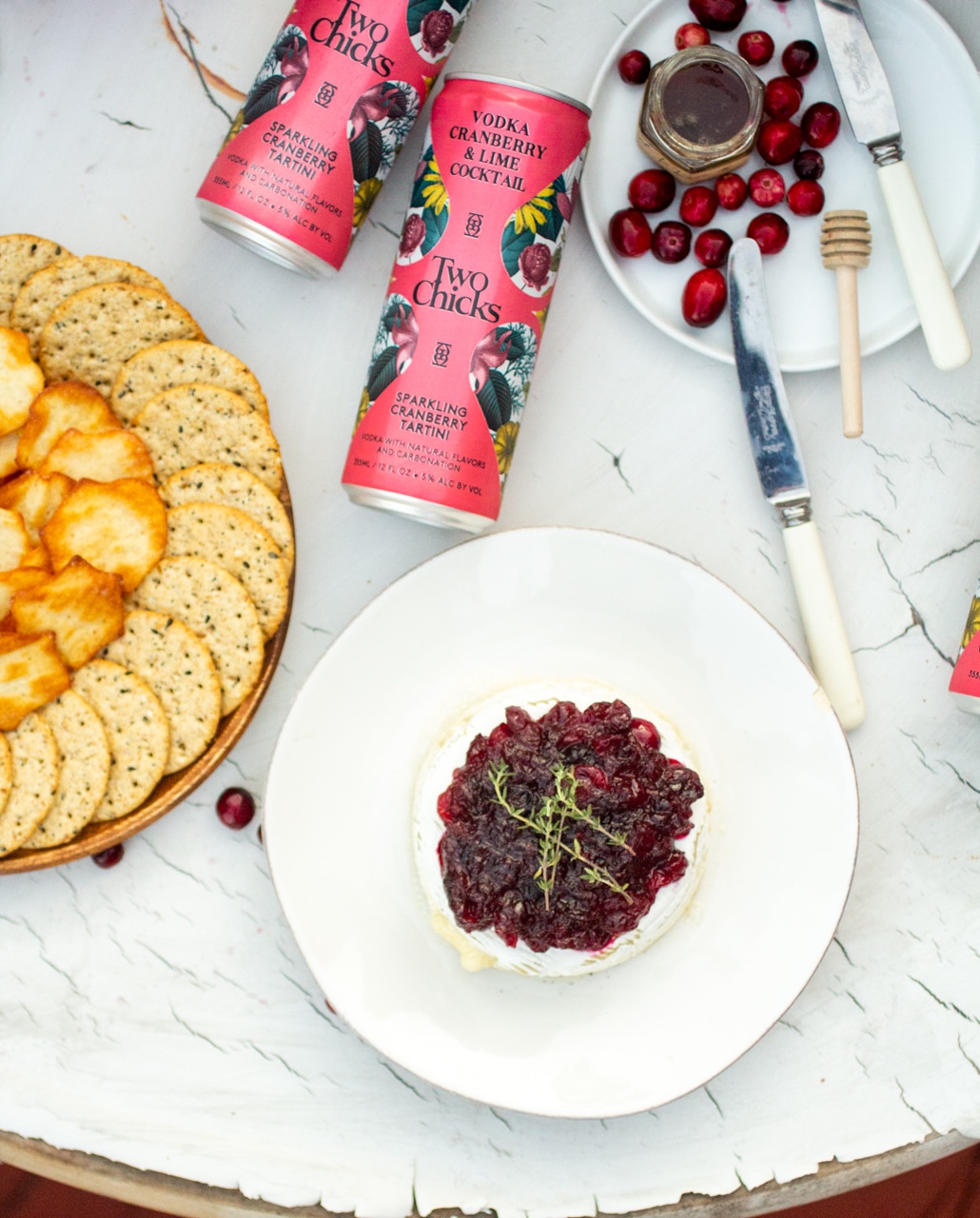 Two cans of Two Chicks on a marble table next to a plate of crackers and a white plate with baked brie.